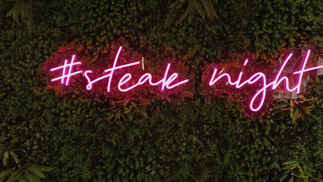 A pink neon sign reading steak night on a plant wall